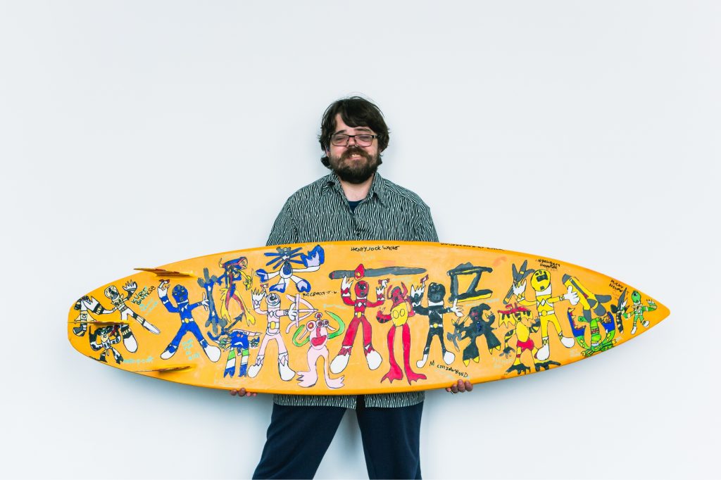 A photo of a bearded man standing and holding a surfboard that has been decorated with drawings of fictional superhero characters