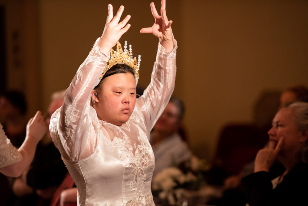A photo of a woman in a wedding dress and tiara raises her hands above her head as an audience watches on.