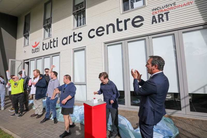 A photo of a group of people cheering and applauding as they stand in front of a building which says Tutti Arts Centre