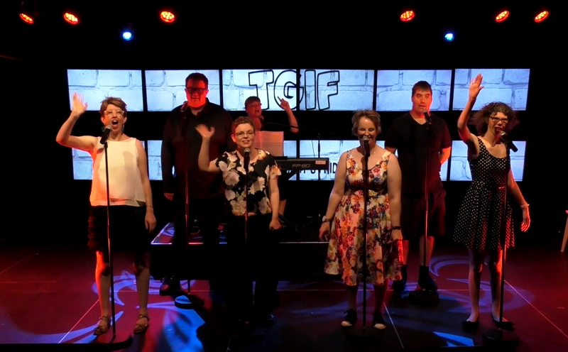 A photo of six singers waving as they stand behind microphones. Behind them is a keyboardist and a screen saying TGIF