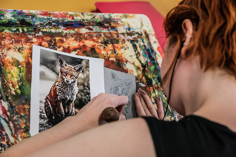 A photo looking over the shoulder of a woman with short red hair as she creates an artwork of a wolf on a paint splattered work stand
