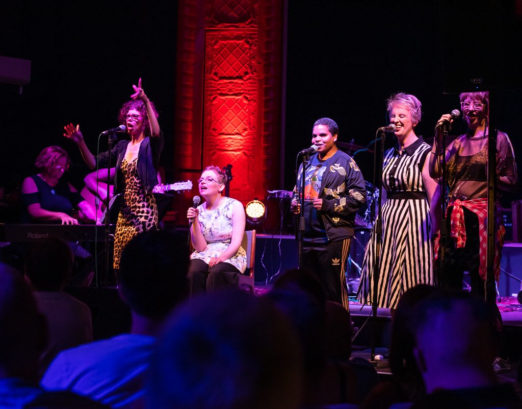 A photo of five women on stage, singing into microphones.