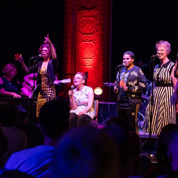 A photo of five women on stage, singing into microphones.