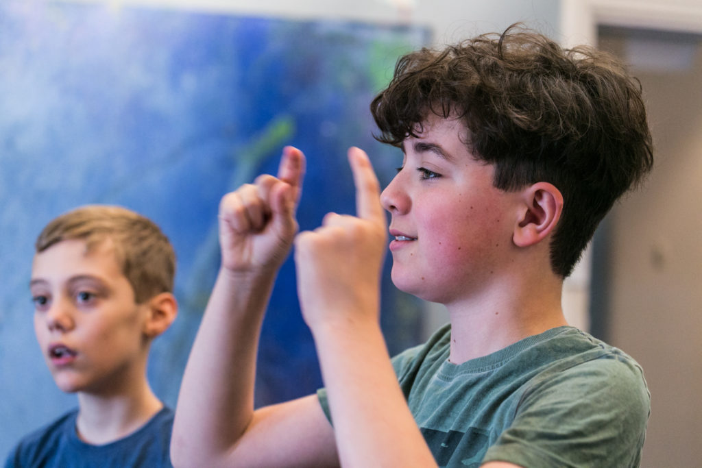 A photo of a boy pointing two fingers upwards with another boy in the background