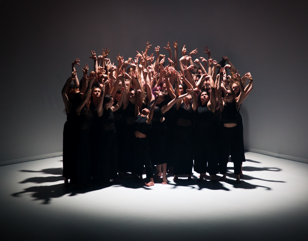 Photo of a large group of dancers raising their hands in the air. The dancers are all dressed in black.