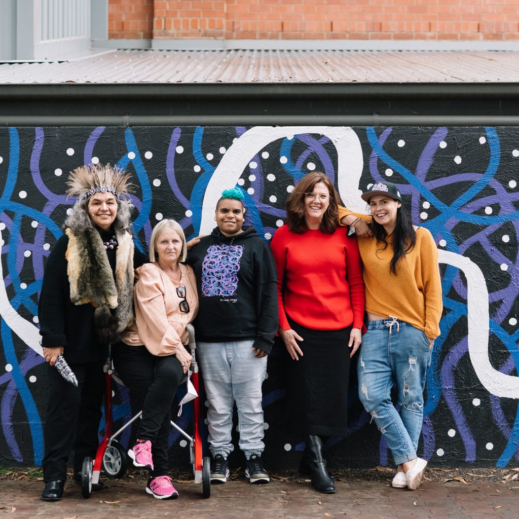 Five people from Tutti, standing in front of a wall with a black, purple, blue and white swirling mural, painted by Jackie Saunders and Elisabeth Close.