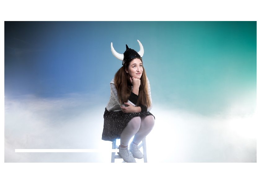 Person sitting on a stool wearing a Viking helmet. They have their head resting on their fist and are smiling and looking to the distance. They are surrounded by a blue stage drop and smoke effects.