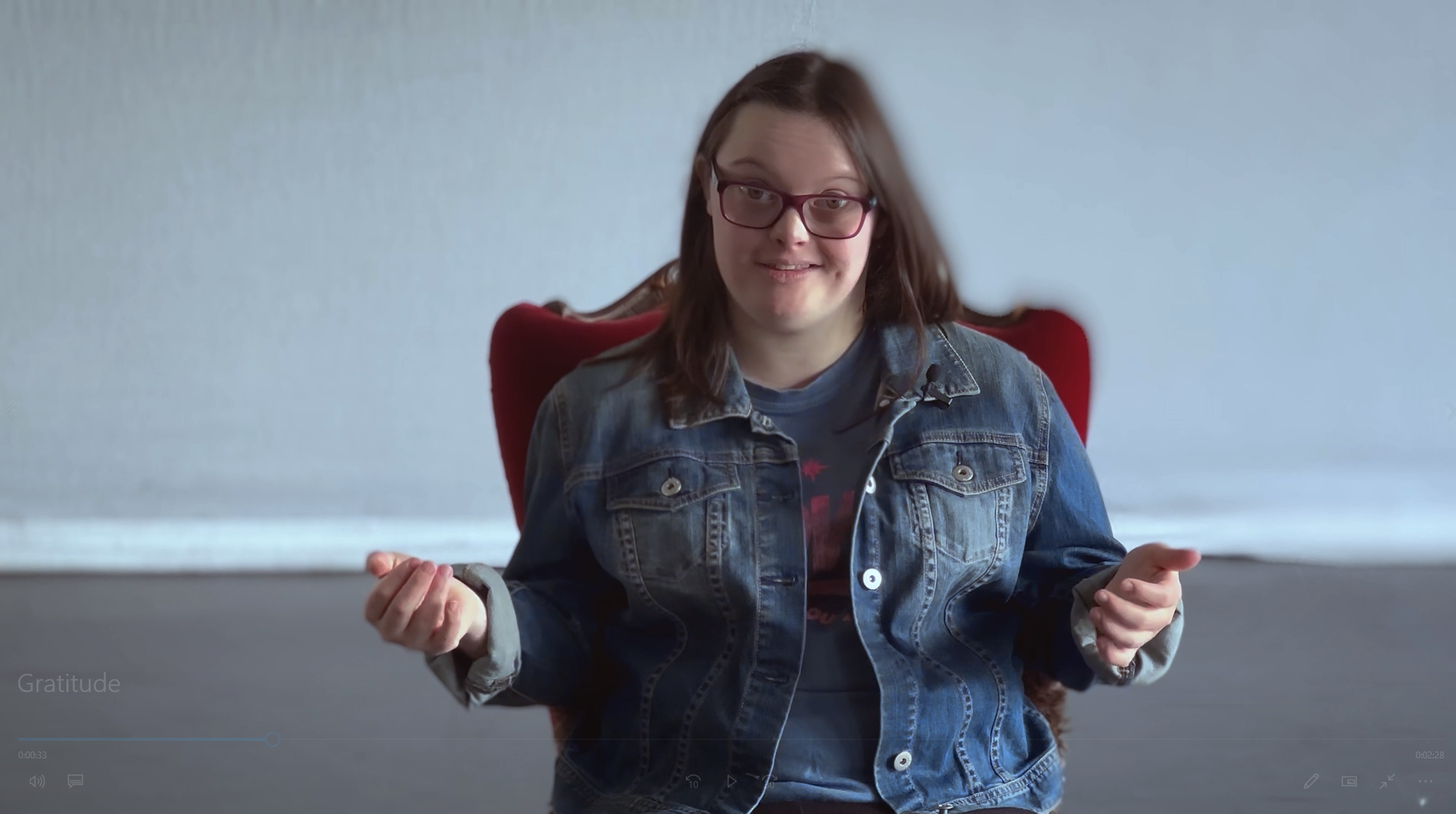 Isi smiling and looking at the camera while sitting on a plush red chair, screenshot from Gratitude video. 