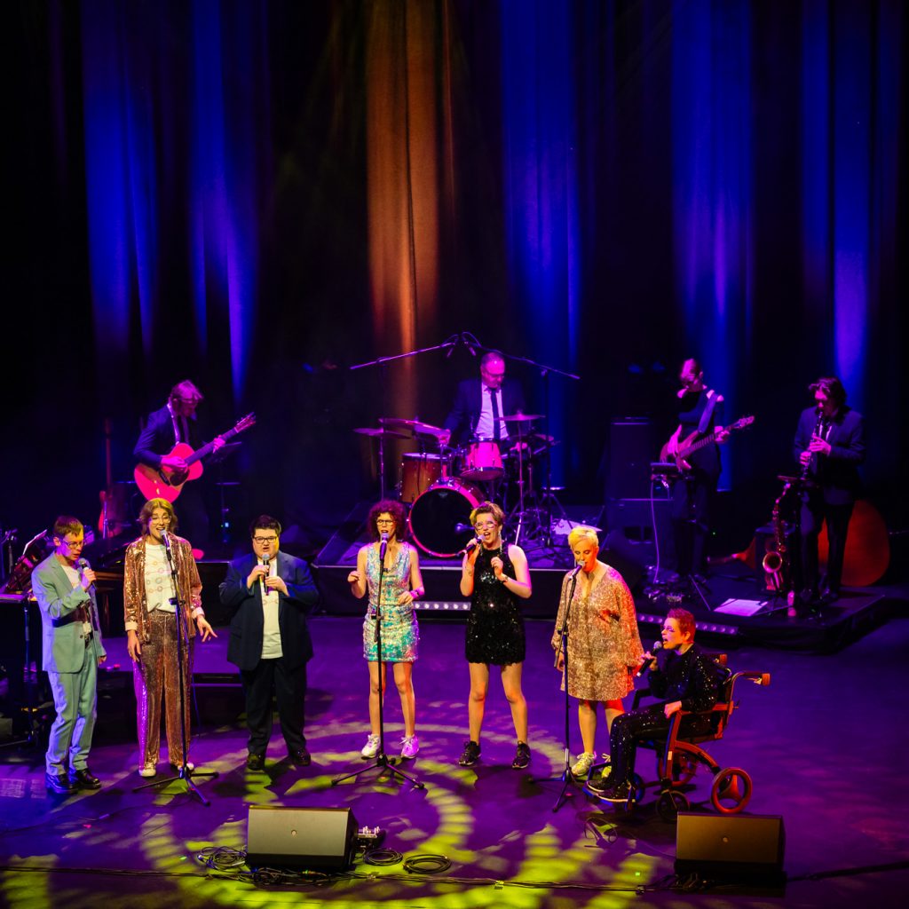 TGIF singing on stage at the Hopgood theatre - there is 7 people on stage wearing a mix of suits and sequined dresses.