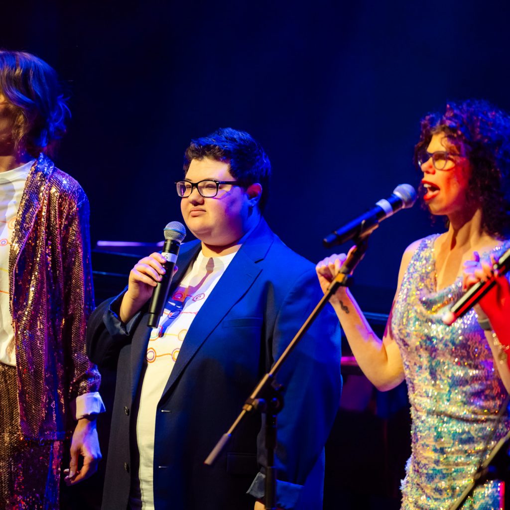 Members of TGIF singing on stage at the Hopgood theatre - there is 4 people in shot, two singing and wearing a mix of suits and sequined dresses.