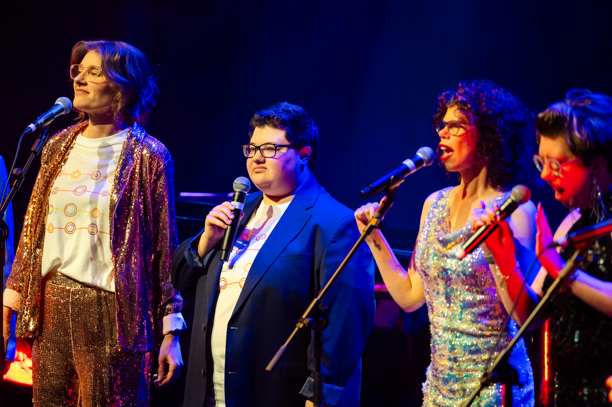 Members of TGIF singing on stage at the Hopgood theatre - there is 4 people in shot, two singing and wearing a mix of suits and sequined dresses.  