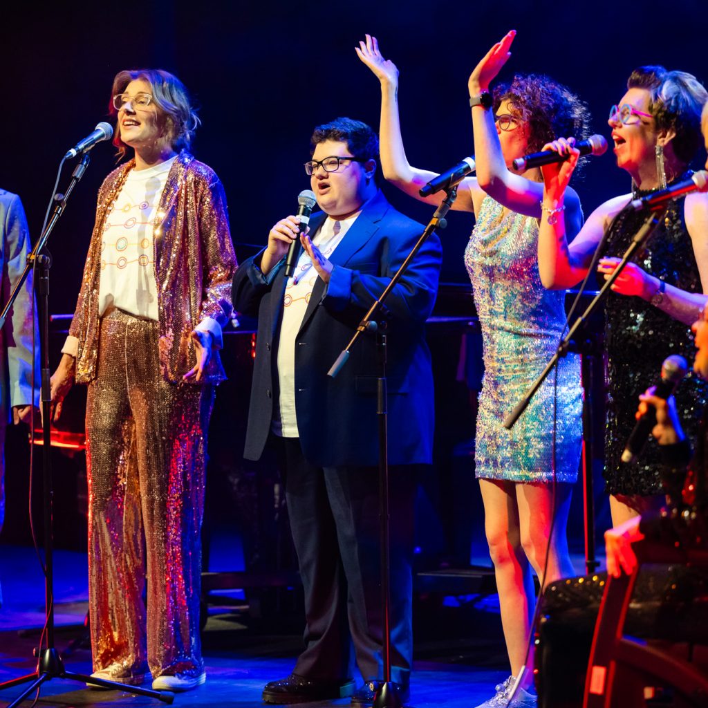 TGIF singing on stage at the Hopgood theatre - there is 7 people on stage wearing a mix of suits and sequined dresses.