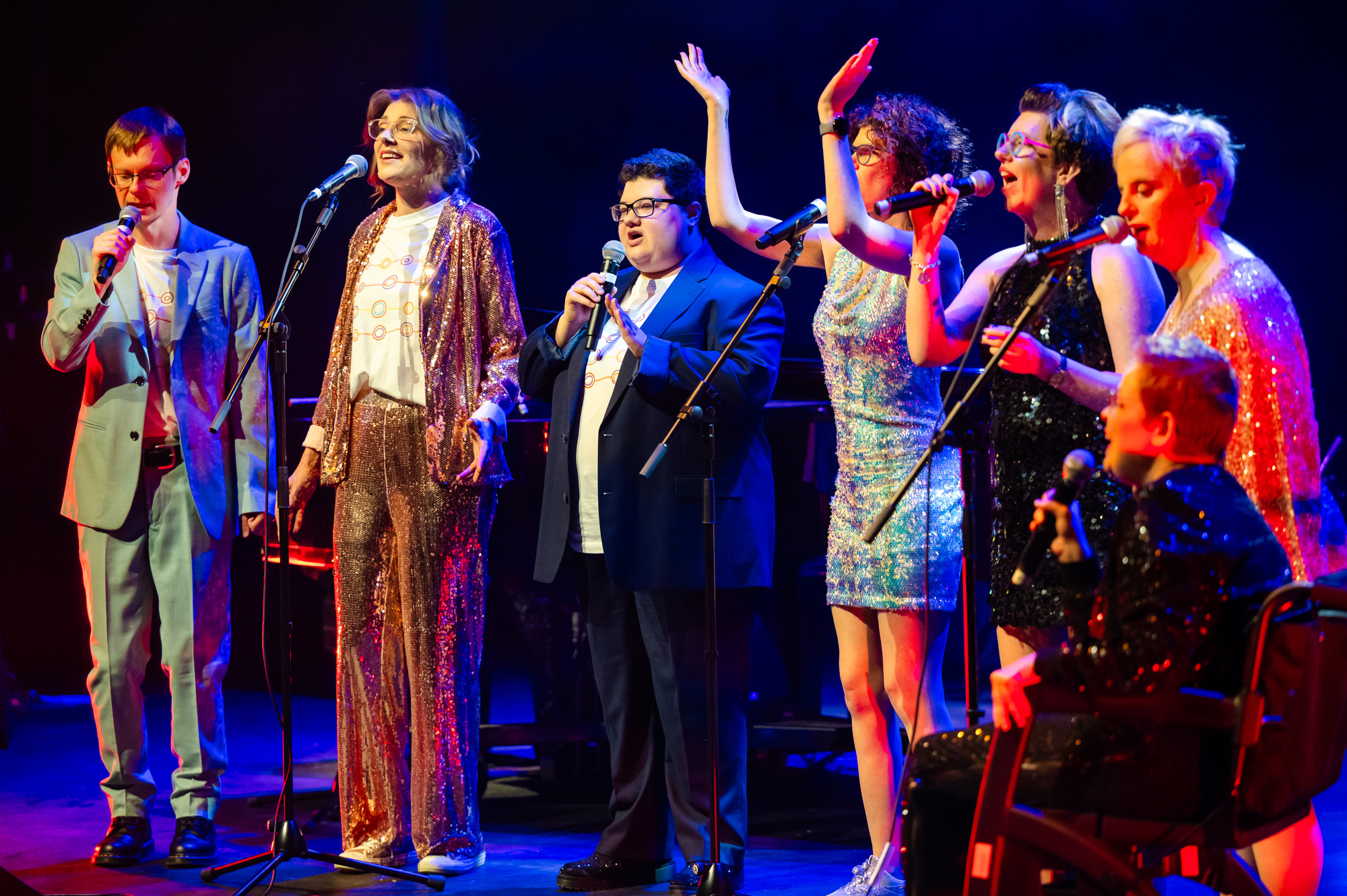 TGIF singing on stage at the Hopgood theatre - there is 7 people on stage wearing a mix of suits and sequined dresses.  