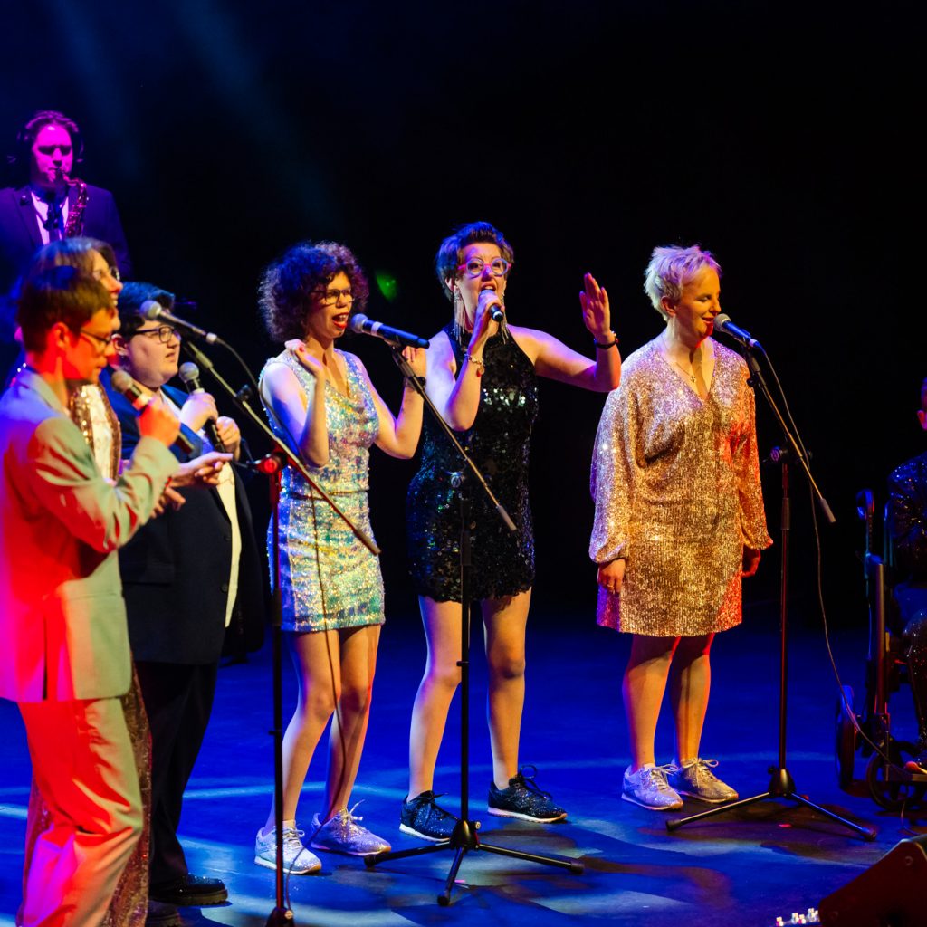 TGIF singing on stage at the Hopgood theatre - there is 7 people on stage wearing a mix of suits and sequined dresses.