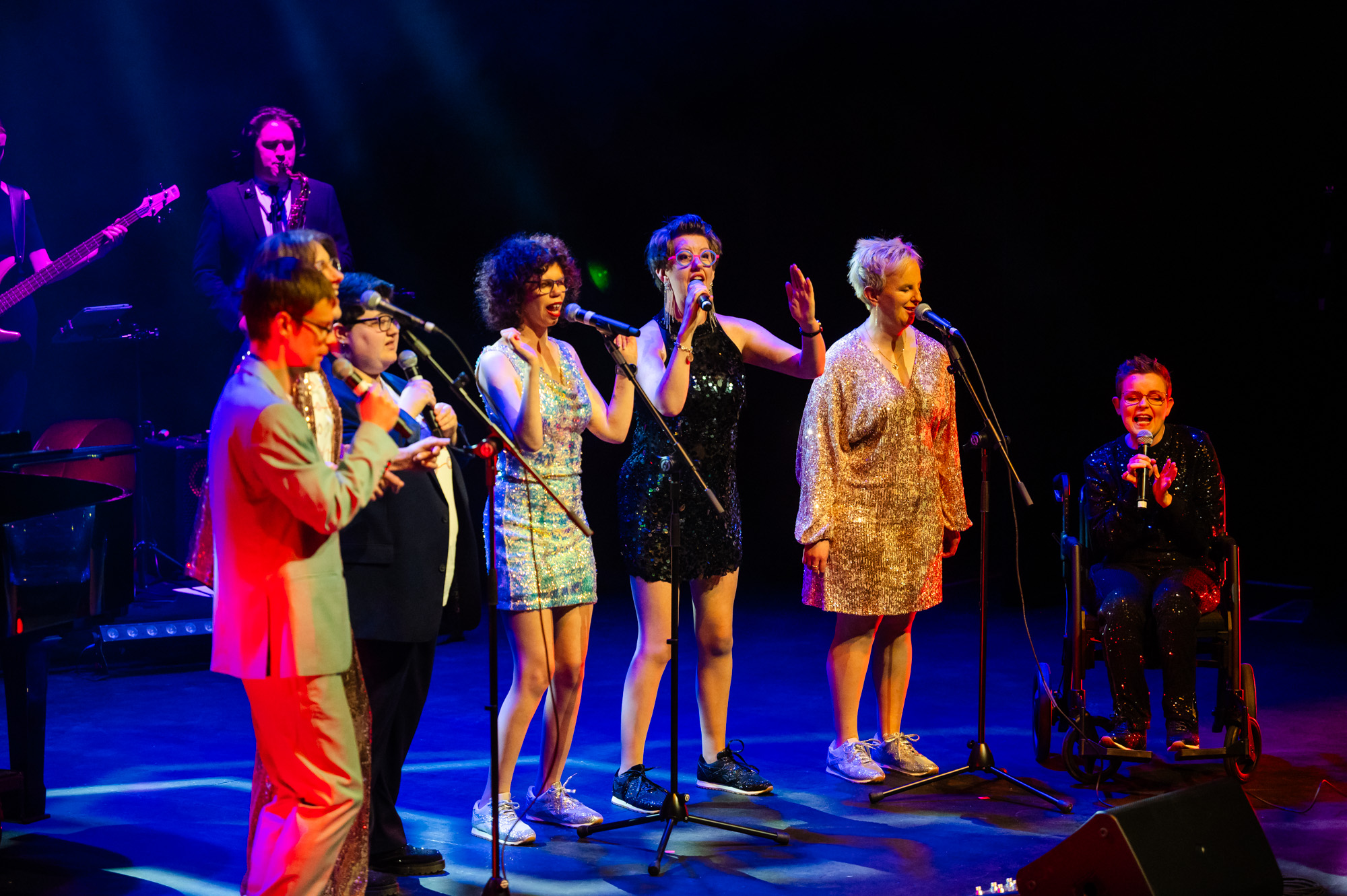 TGIF singing on stage at the Hopgood theatre - there is 7 people on stage wearing a mix of suits and sequined dresses.