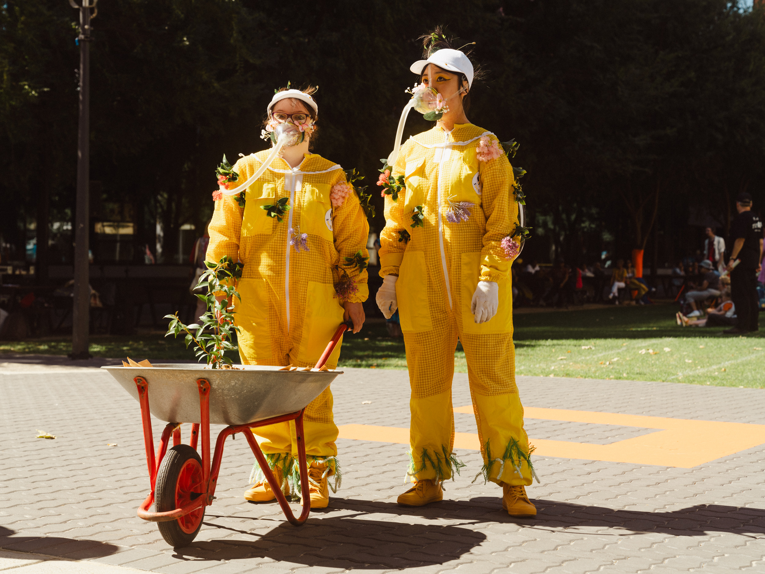 Annalise and Isi dressed in bright yellow beekeeper costumes. Thet are wearing oxygen masks, are covered in flowers and pushing a wheelbarrow. 