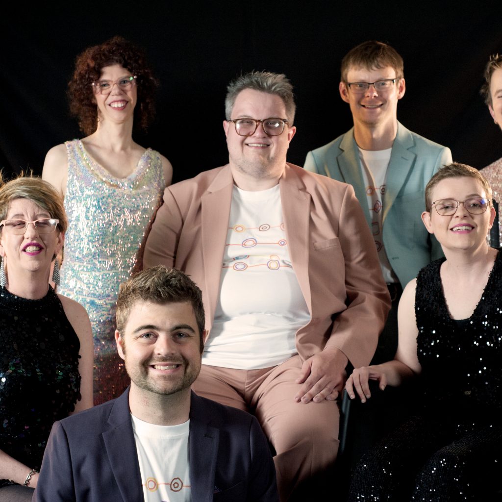 Professional photo shot of TGIF - some members standing and others sitting and one member is kneeling down in front, they are forming a group and all smiling at the camera.
