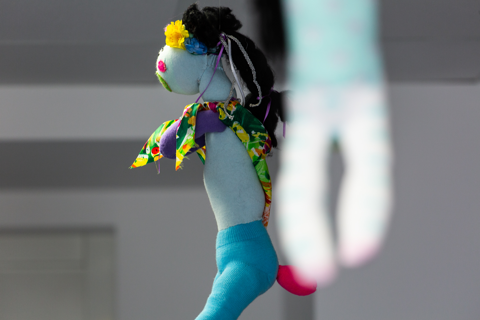 A knited sculpture of a doll inspired by Frida Kahlo handing from the ceiling. It has long black hair, a flower crown, a brightly coloured scarf, blue legins with a pink tail poking out.