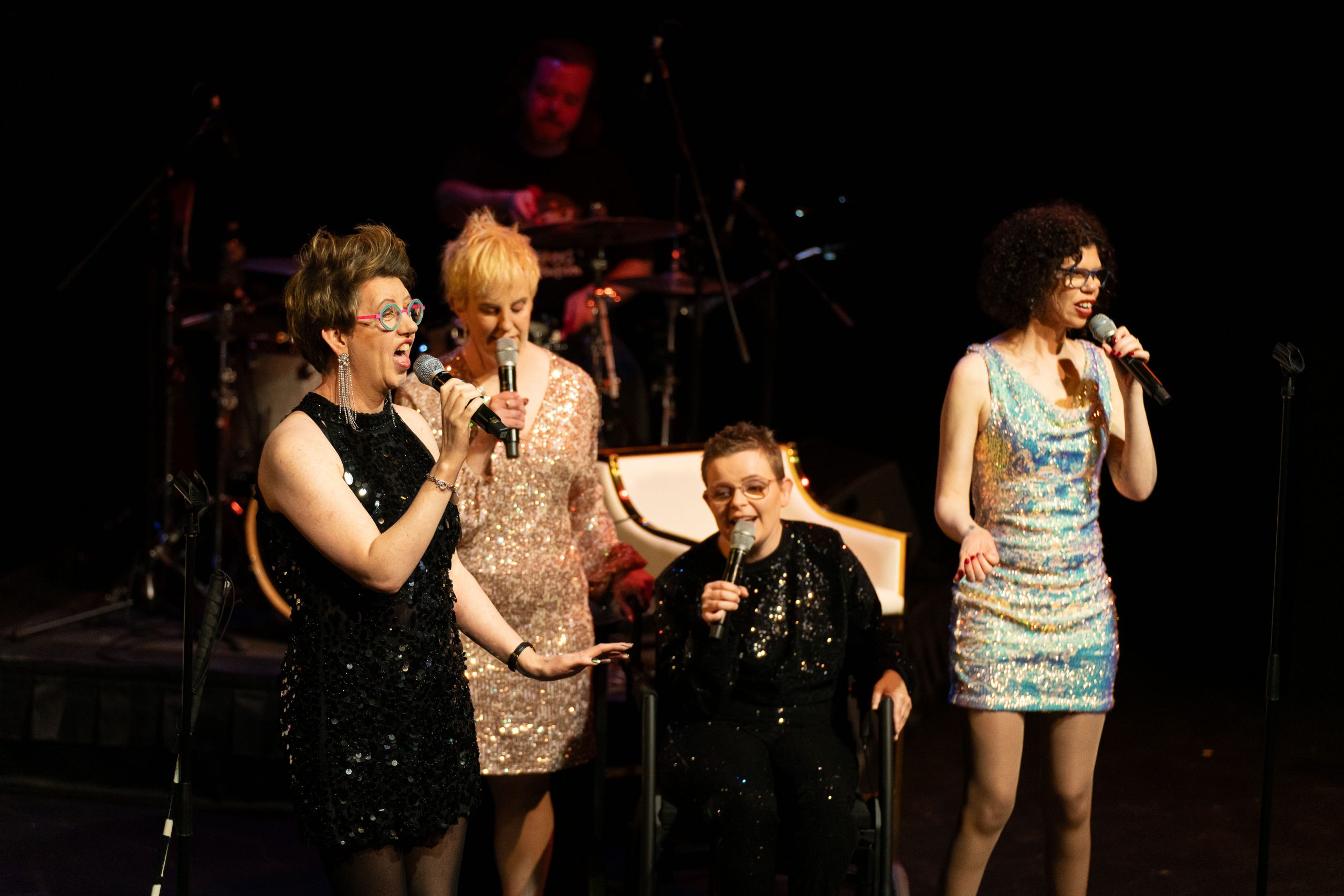 The four members of The Sisters of Invention performing on stage, they are all singing into microphones. 