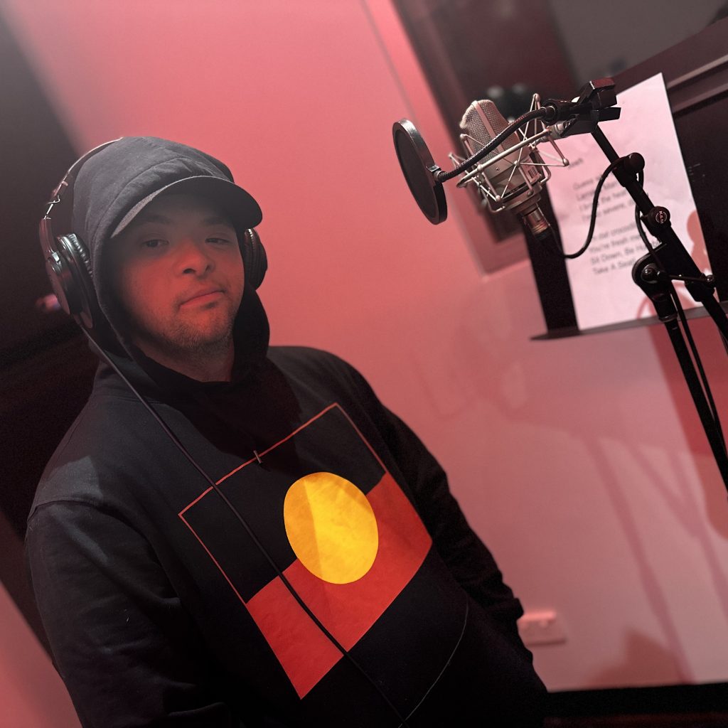 A person wearing a hoodie featuring an Aboriginal flag, in red lighting in a recording booth complete with headphones and a microphone.