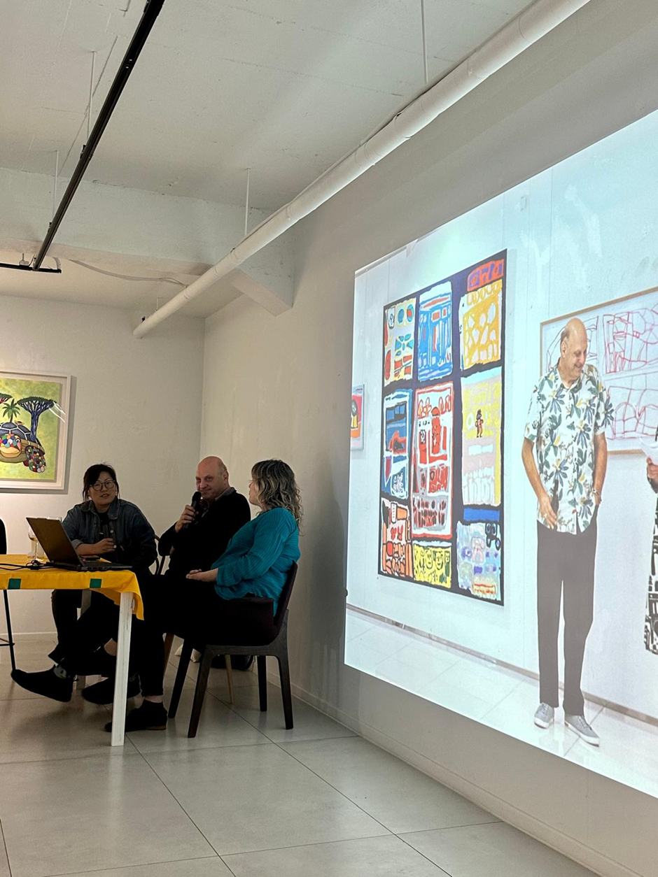 Lewis sitting at a desk in front of a project screen showing him by his artworks - he is talking into a microphone. 