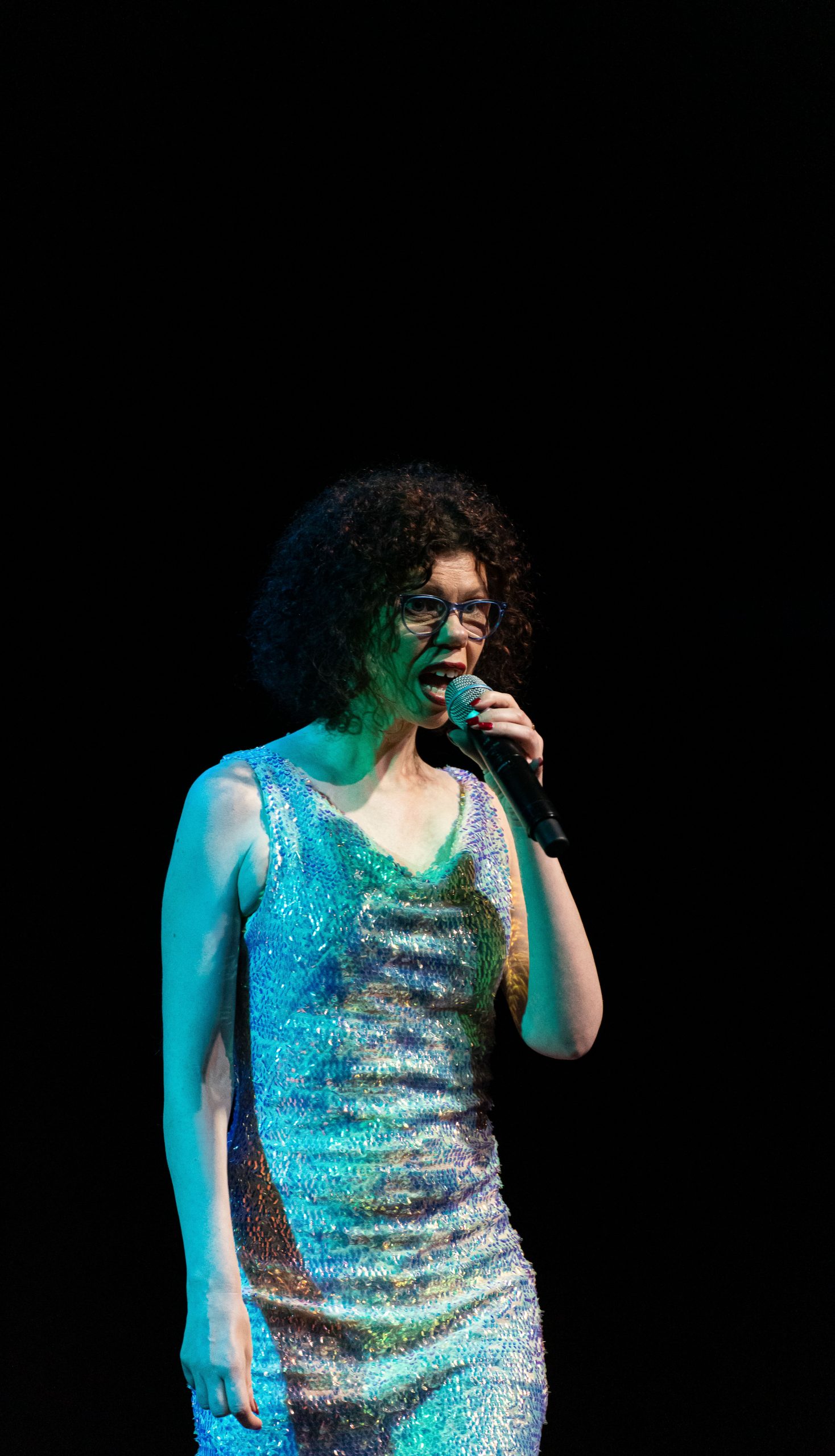 AImee singing into a microphone on a dark stage with a sequin dress on. 