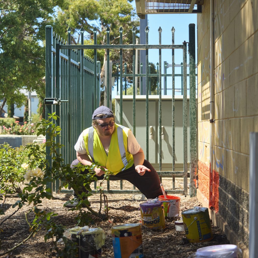Jacko Hooper squatting down and pointing at the signal hut - he's surrounded by open tins of paint.