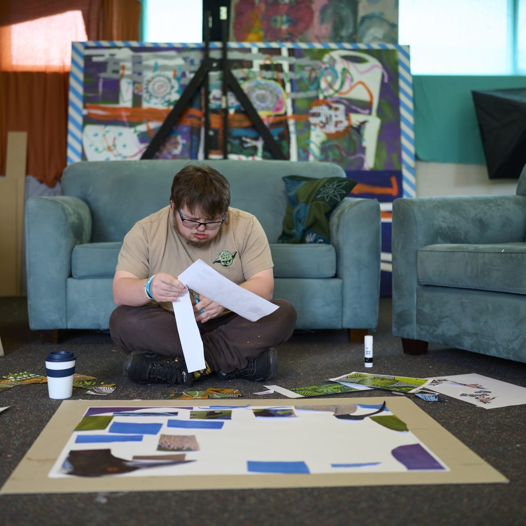 Person cutting out printed copies of different artworks and arranging them on a large collage board on the ground.