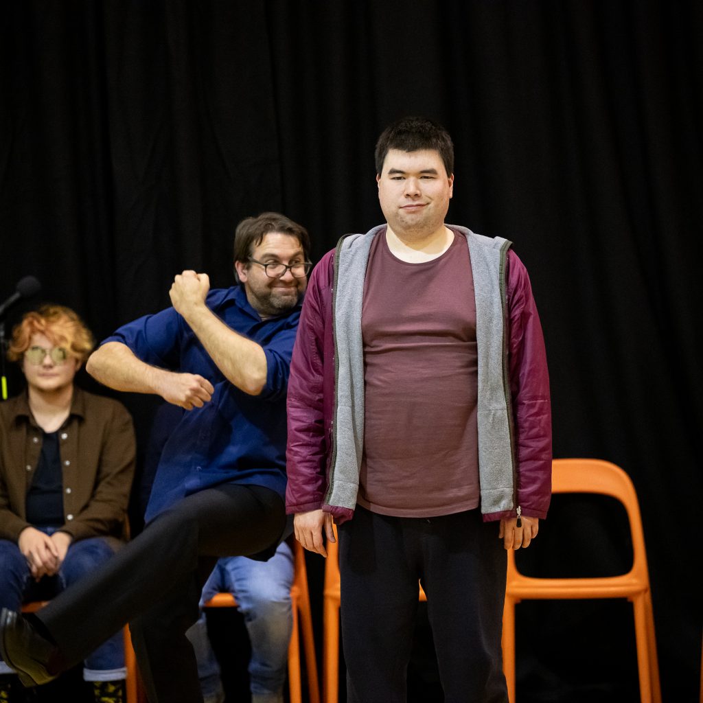 a man stands still looking at the camera, another man stands behind him pulling a gesture with his arm.