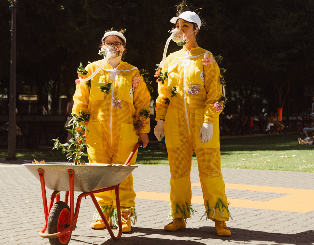 Two people stand dressed in yellow beekeeping suits covered in flowers, one holds a wheelbarrow with a plant sticking out of it.